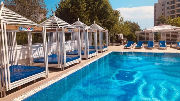 Una piscina con tumbonas y gazebos, rodeada de árboles y un edificio al fondo.