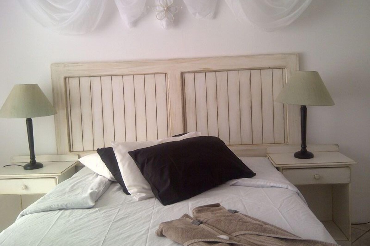 A bed with white bedding, two black pillows, and a pair of brown robes on the side. There are two bedside tables with lamps.