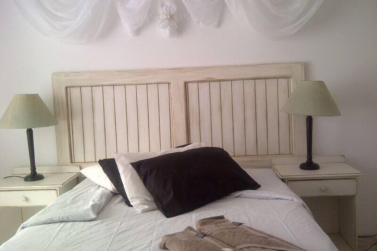 Un lit avec une literie blanche, deux oreillers noirs et une paire de peignoirs marron posés sur une chaise à proximité. Il y a deux tables de chevet avec des lampes.