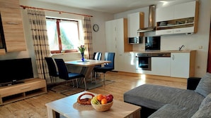 A modern living room with a TV, sofa, and dining area with chairs and a table. A kitchen with appliances and cabinets is visible in the background. - Schmuckenhof (Marktschellenberg)