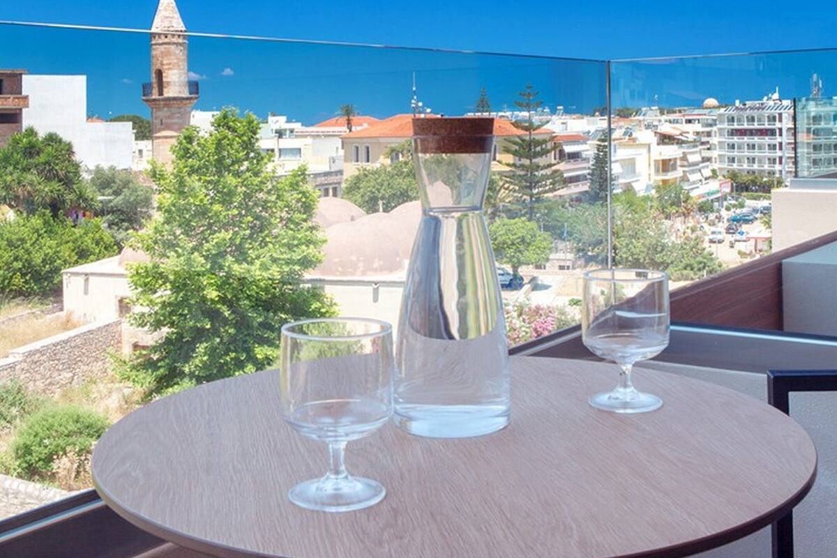 A round table with two wine glasses and a clear glass pitcher on a balcony overlooking a cityscape with buildings and a clear blue sky.