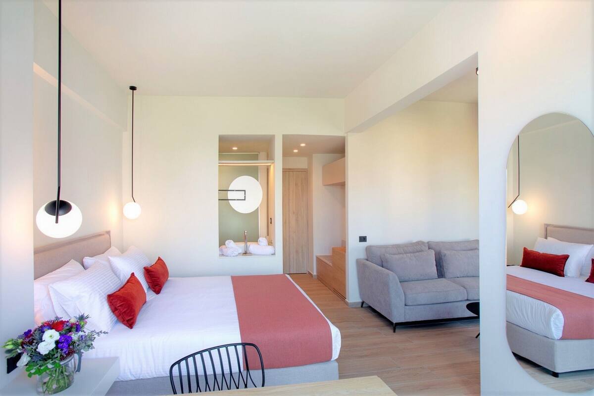 A modern hotel room with a large bed, a sofa, a dining table, and a mirror.