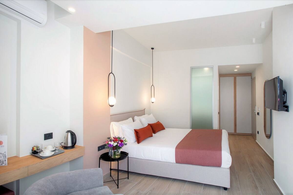 A modern hotel room with a large bed, bedside table with a kettle, a small table with flowers, and a TV mounted on the wall.