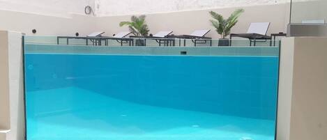 Una amplia piscina interior con agua cristalina y azul, rodeada de sillones de descanso y pequeñas palmeras.