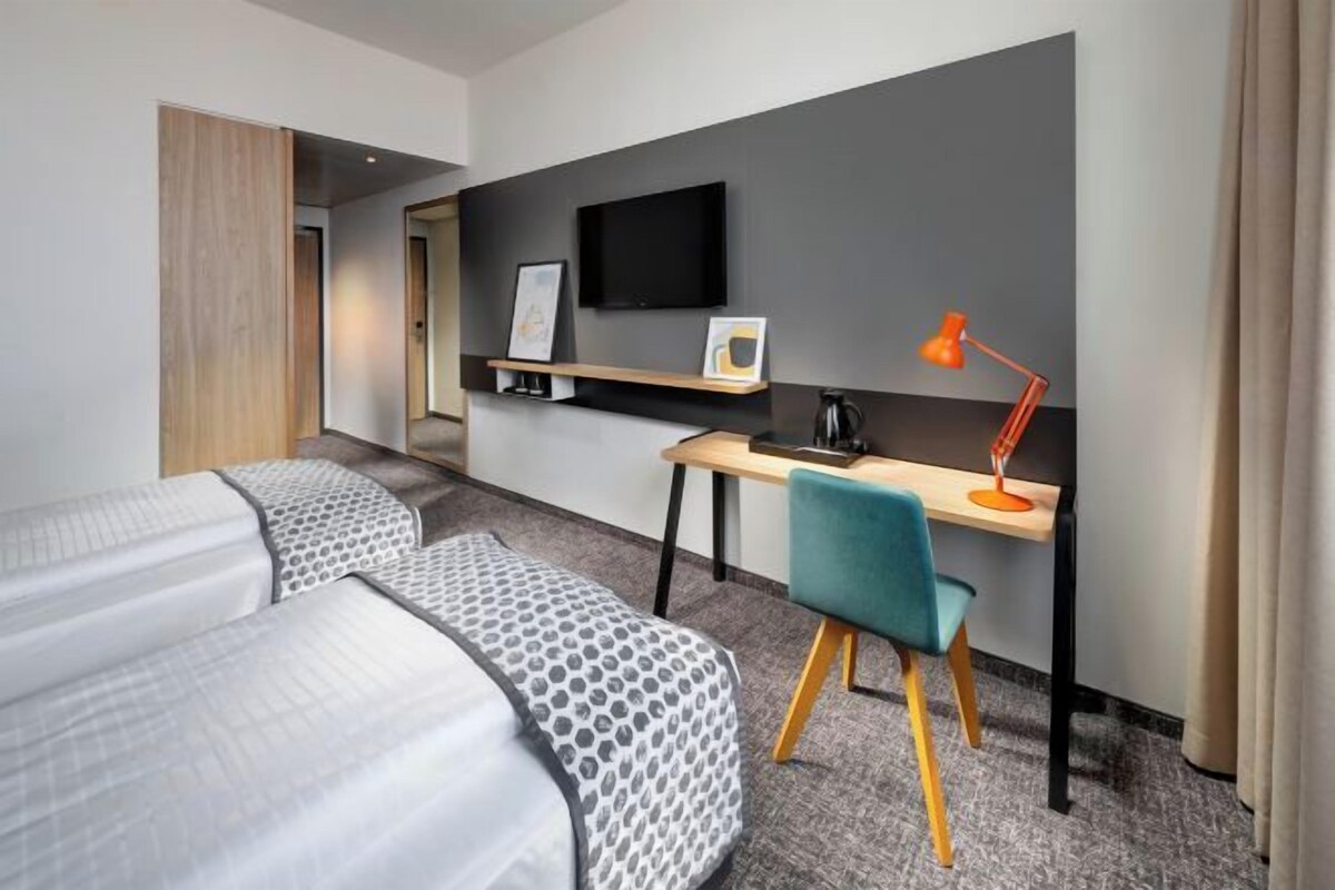 Ett hotellrum med en säng, ett skrivbord, en stol, en tv och en kaffebryggare.