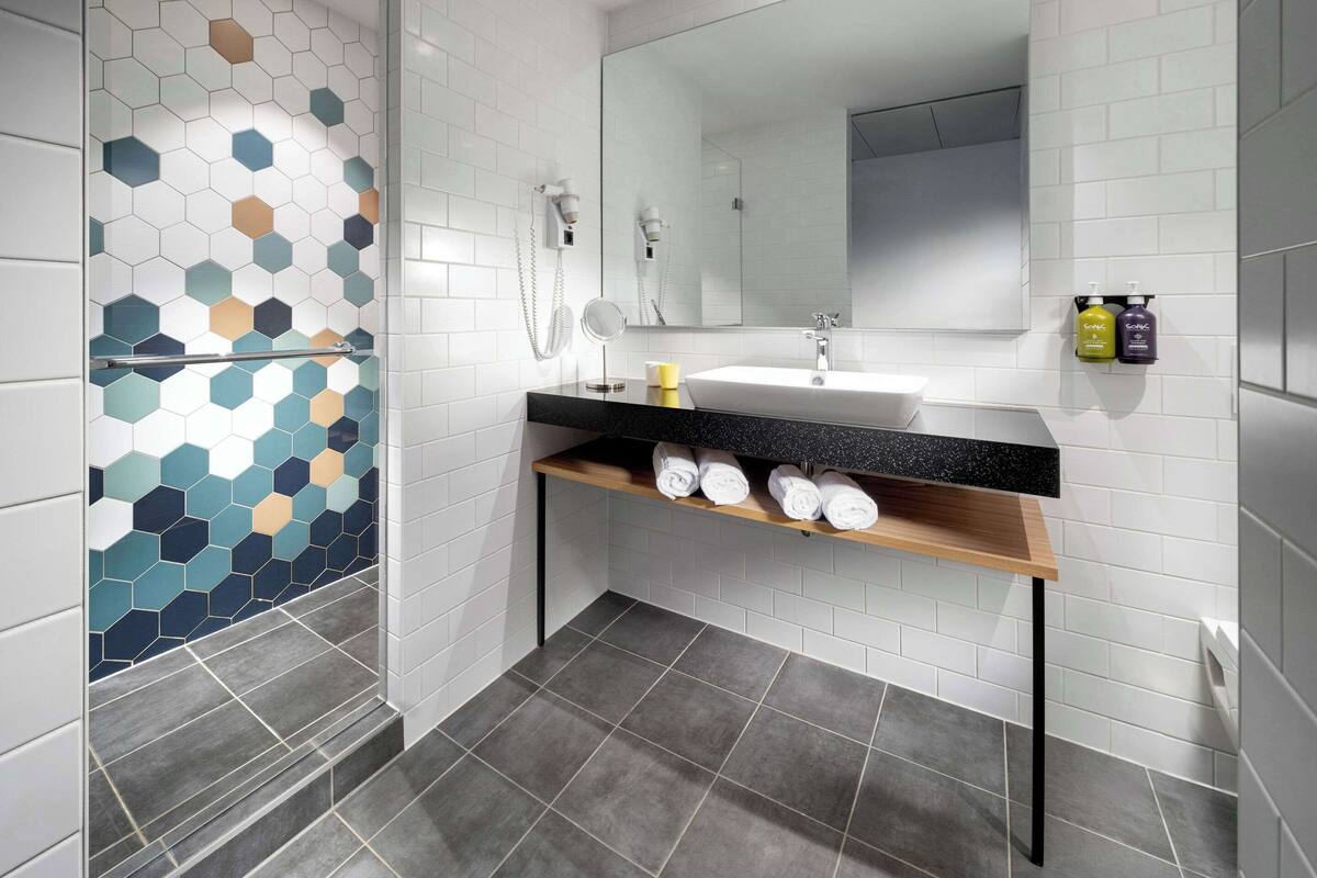 Un baño moderno con pared de azulejos blancos, encimera negra, espejo y estante con toallas.