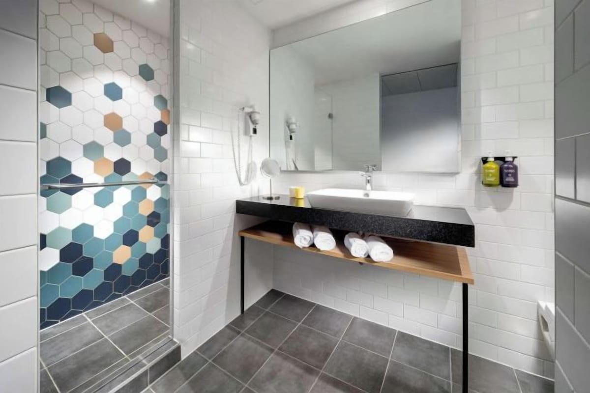 Um banheiro moderno com painel de azulejos hexagonais, bancada preta, espelho e prateleira com toalhas.