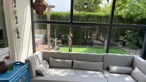 A modern living room with a beige sectional sofa, a blue side table, and a large window offering a view of a well-maintained garden. - Bungelow "Duinvogel" Beach with bath in the garden (Noordwijk)