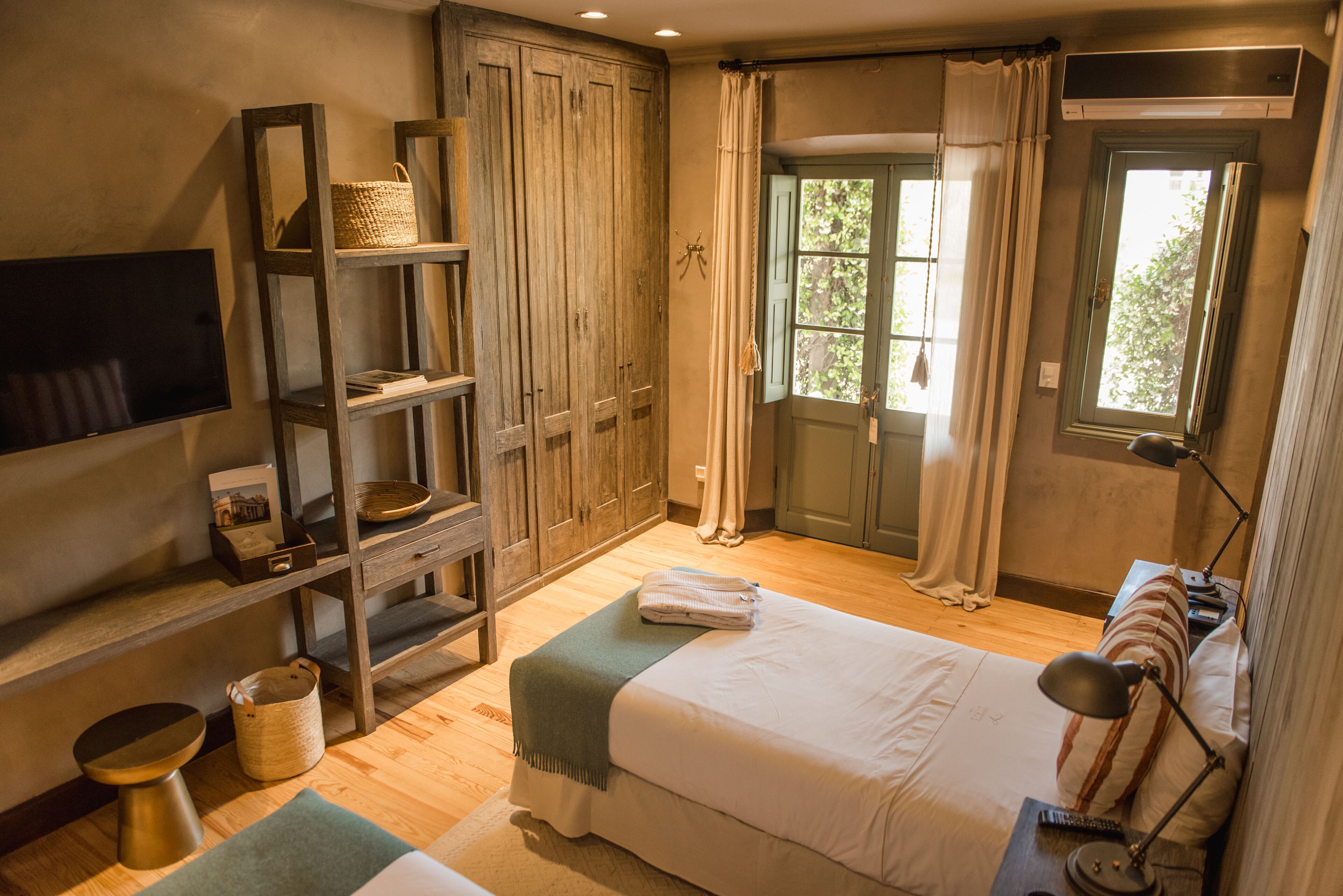 Un dormitorio con cama, mesita de noche, lámpara, estante con cesta y ventana con cortinas.