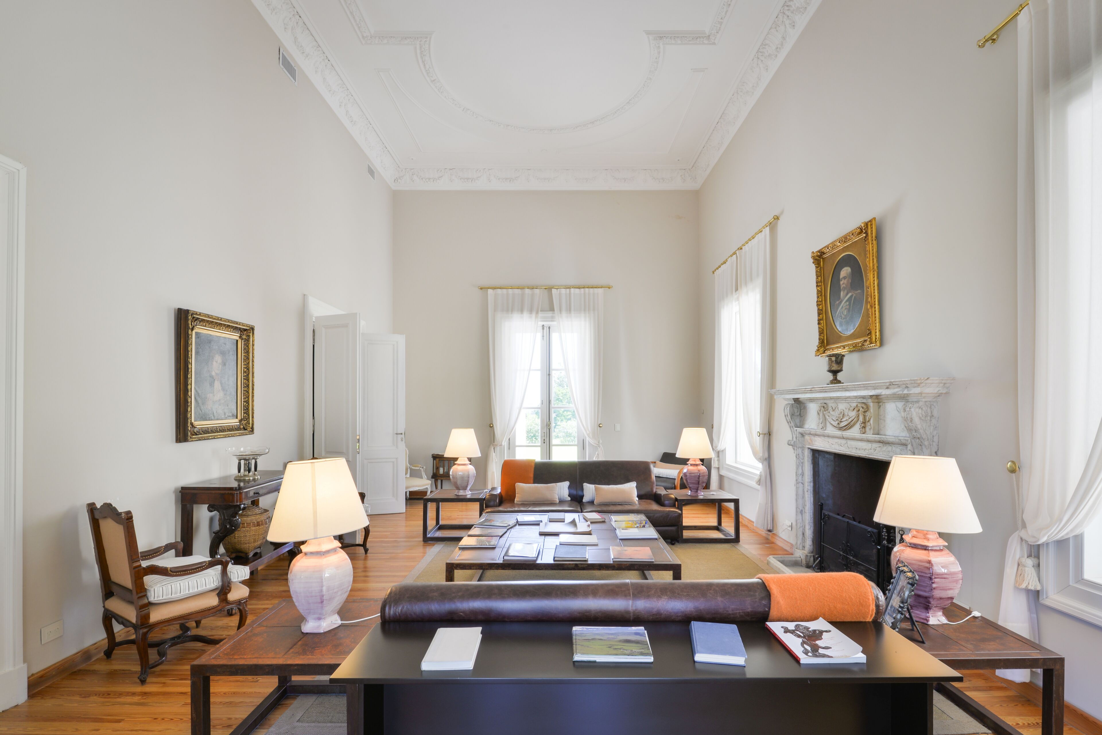 Een ruime kamer met een grote centrale tafel, twee banken, een open haard en kunstwerken in lijsten aan de wanden.