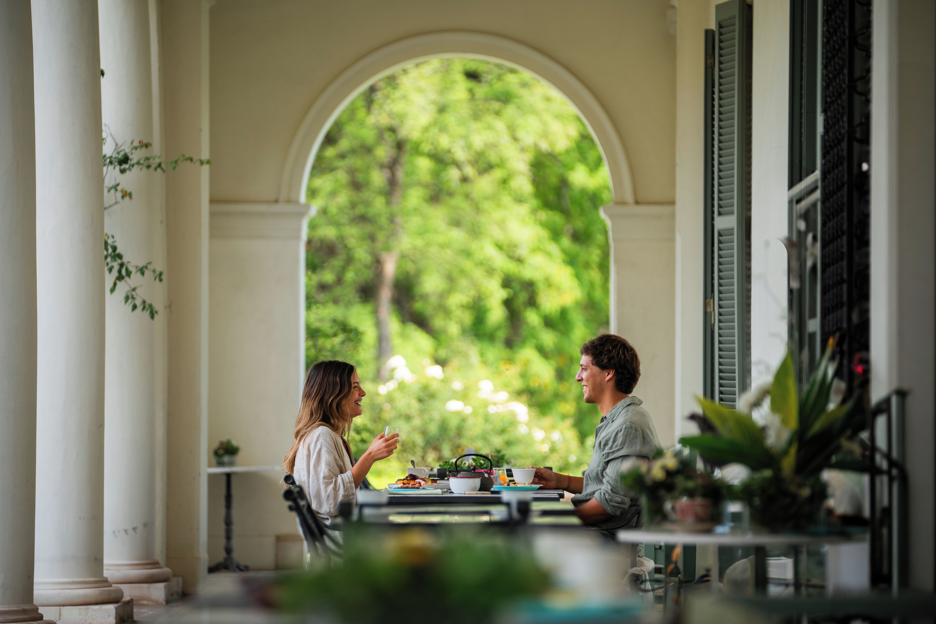 A dining area with a table set for two, surrounded by columns and a view of lush greenery.