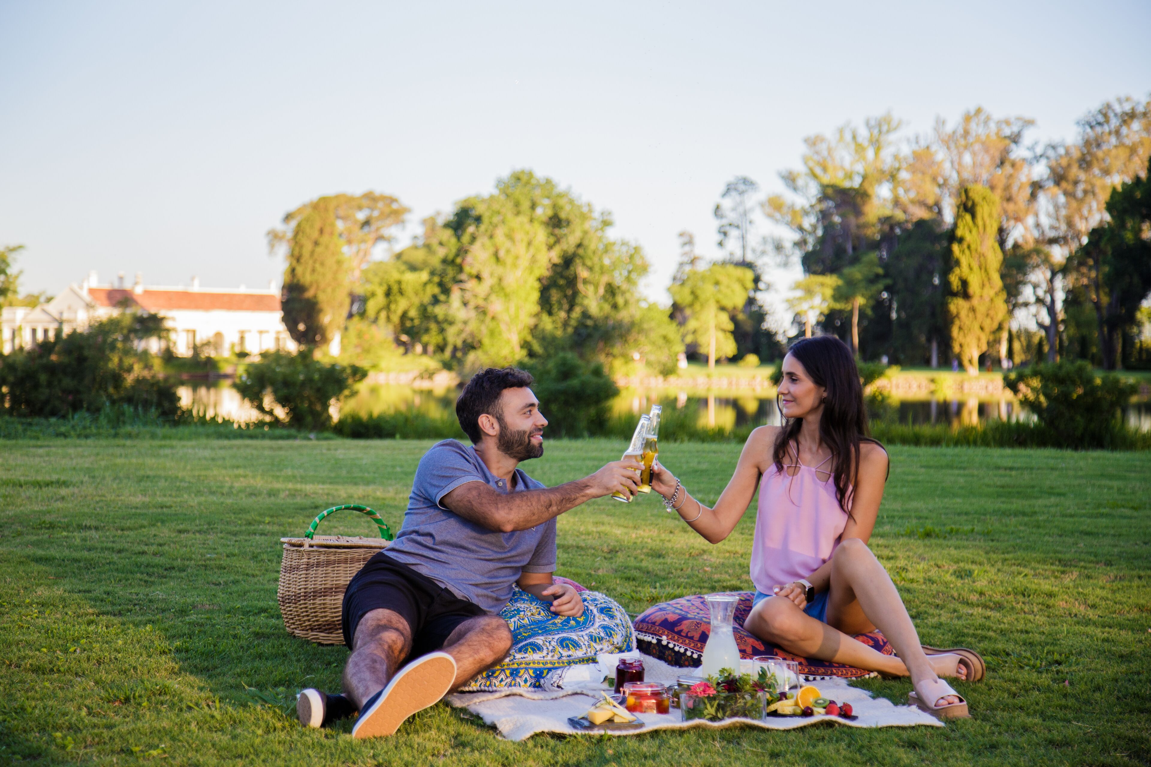 A couple sitting on a blanket in a park, enjoying a picnic with food and drinks.