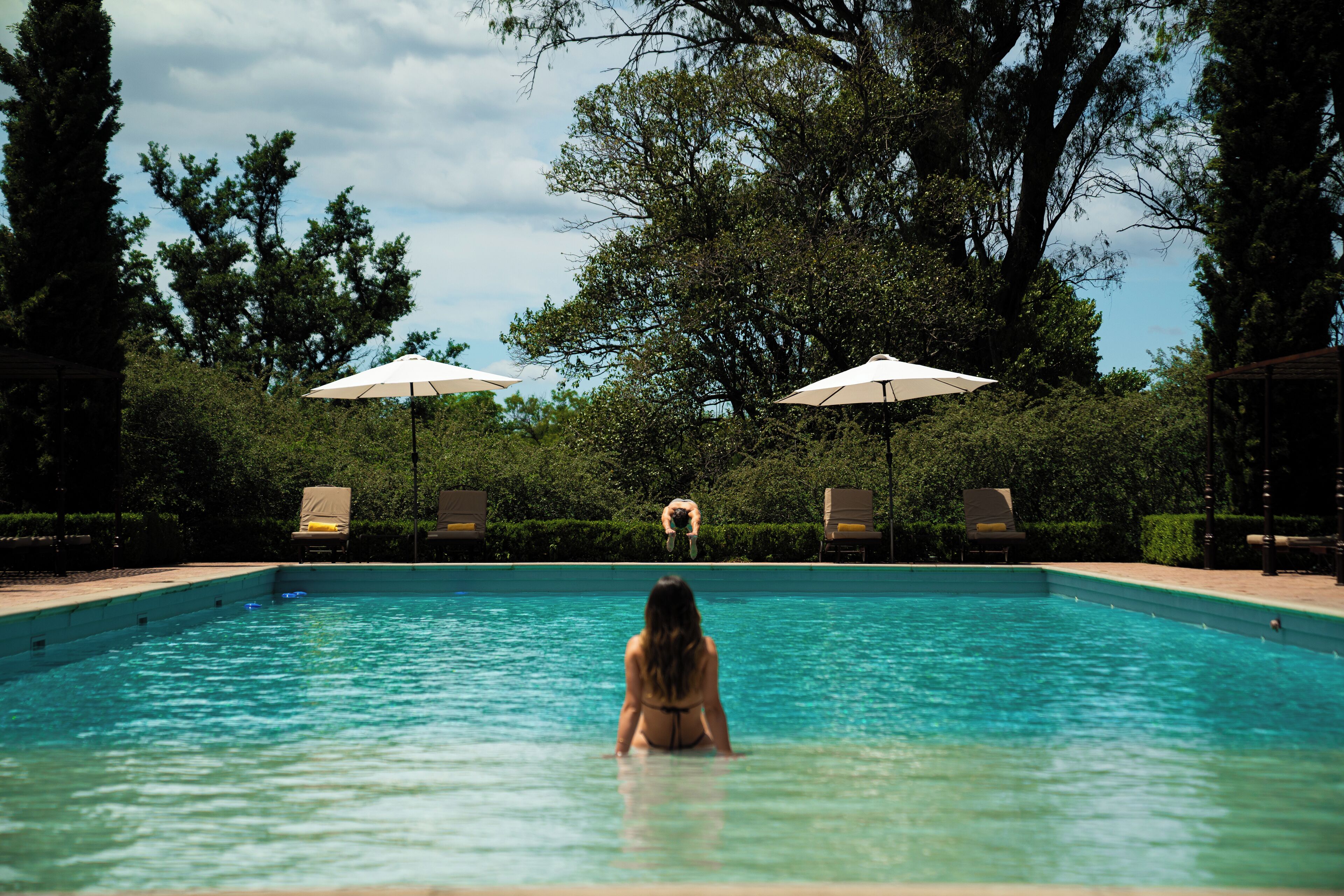 A pool with a person in the water, surrounded by trees and umbrellas.
