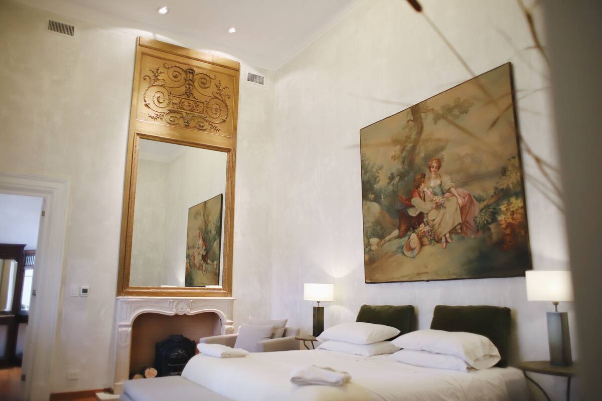 Ett hotellrum med en stor säng, en öppen spis, en stor spegel och en tavla på väggen.