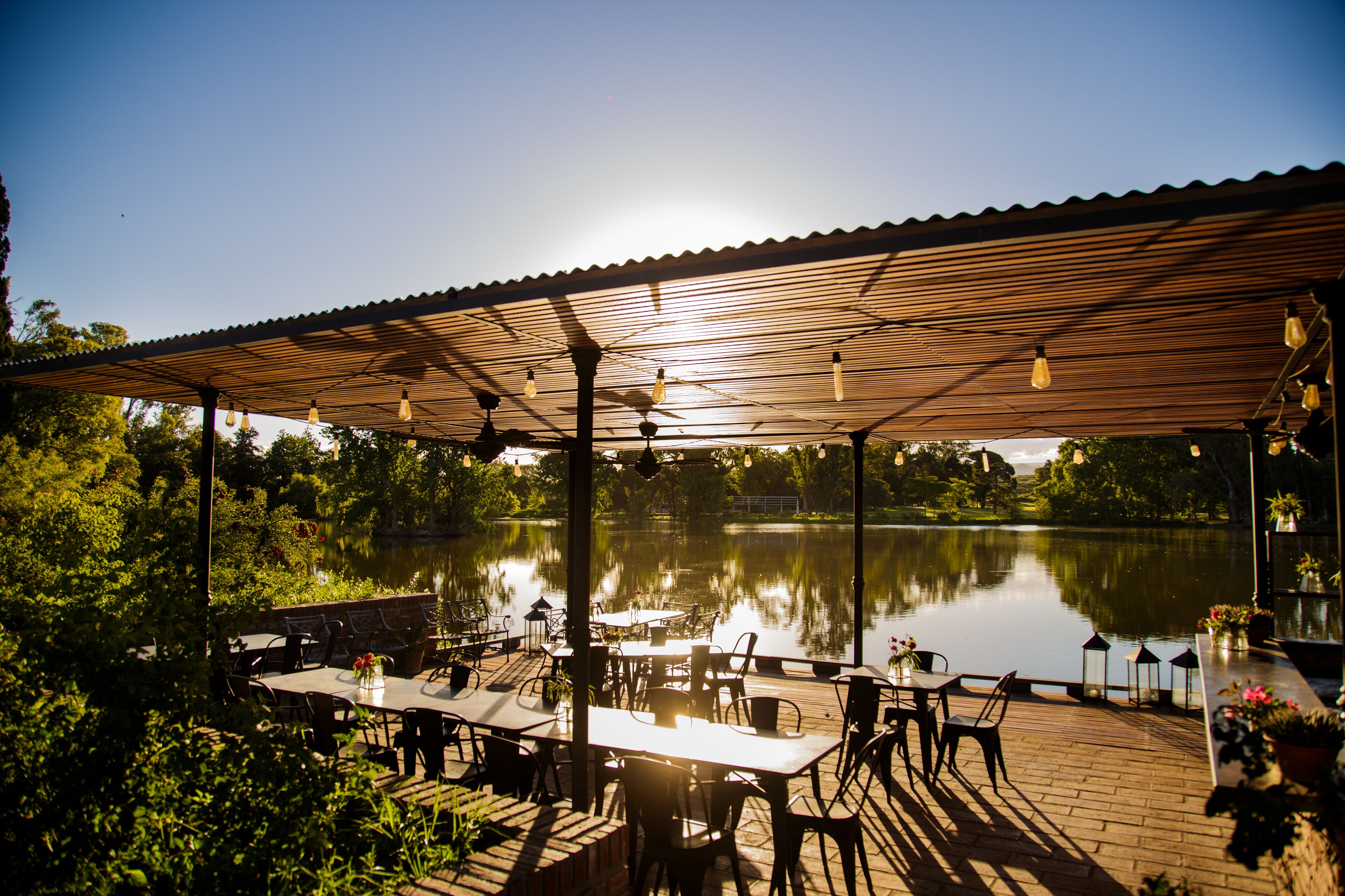 An outdoor dining area with tables and chairs, a wooden pergola, and a view of a lake.