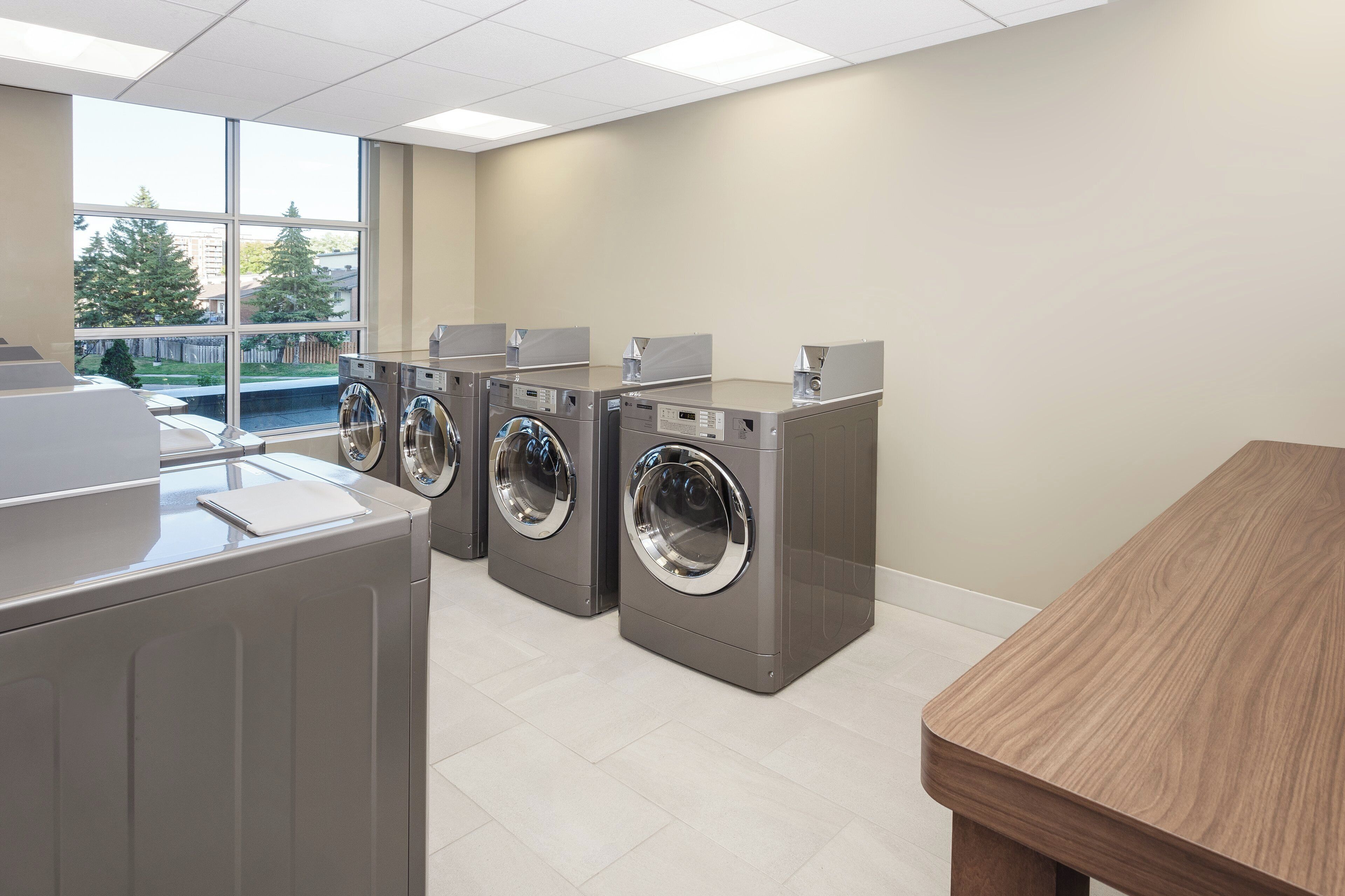 A laundry facility with four front-loading washing machines, a wooden table, and a view of a residential area through large windows.