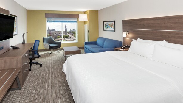 A hotel room with a large bed, a desk, a chair, a sofa, a TV, and a window with a city view. - Holiday Inn Express & Suites Downtown Ottawa East by IHG (Ottawa)