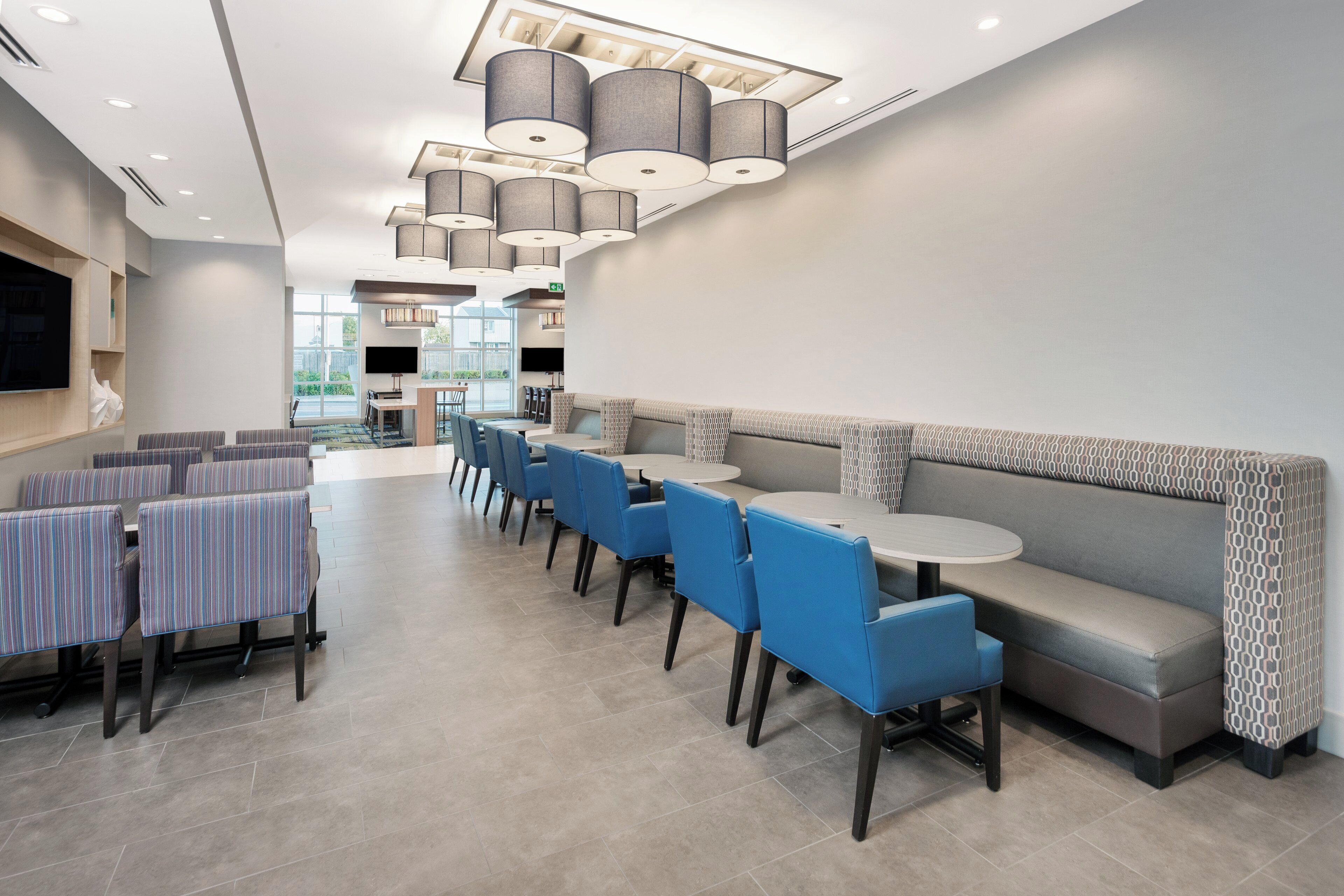 A modern dining area with a mix of seating options including booths and tables, a flat-screen TV mounted on the wall, and a ceiling with recessed lighting and pendant lamps.