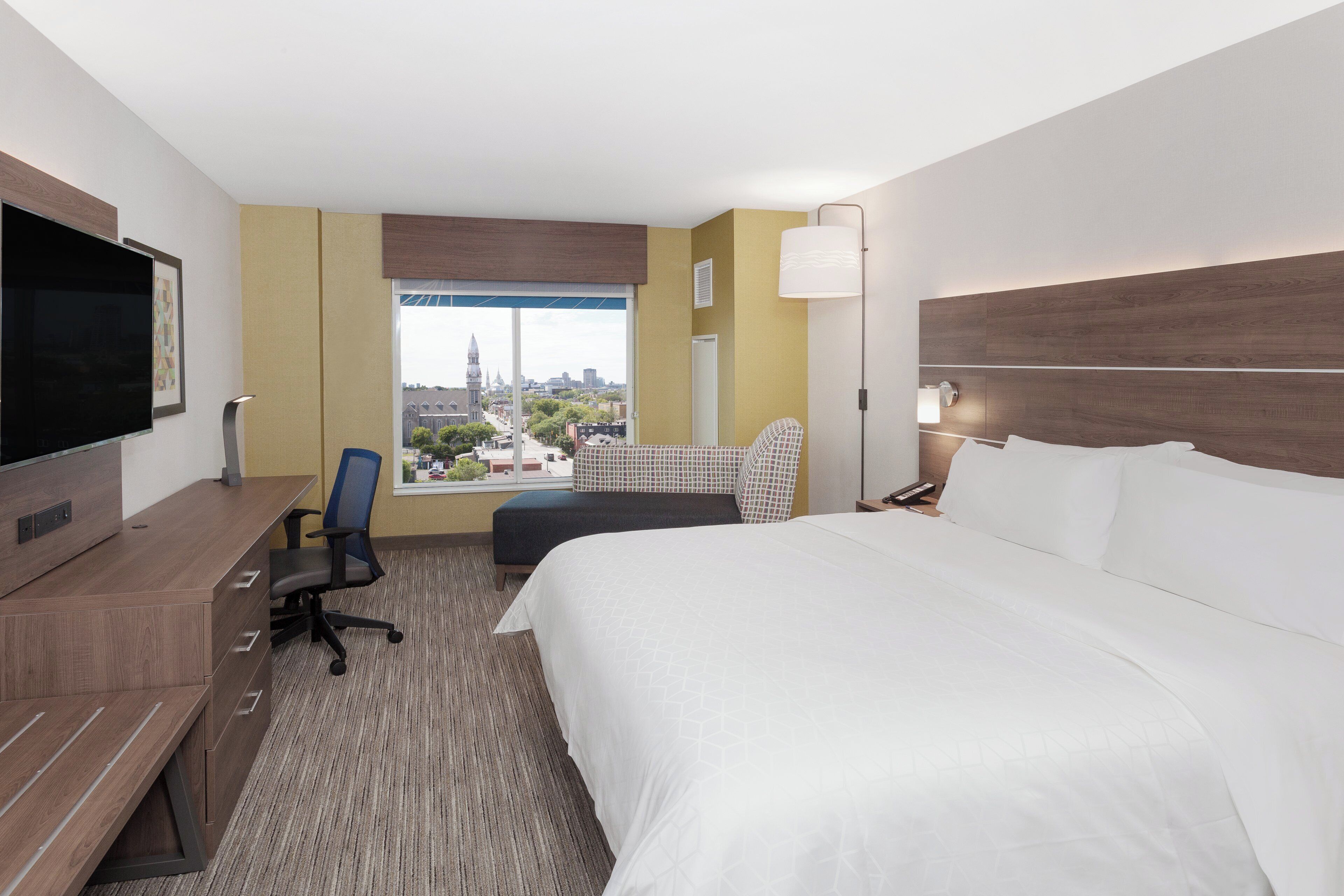 A hotel room with a large bed, a desk with a chair, a television, a window with a city view, and a lamp.