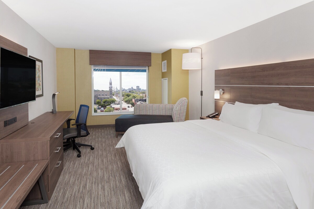 A hotel room with a large bed, a desk with a chair, a television, a window with a city view, and a lamp.