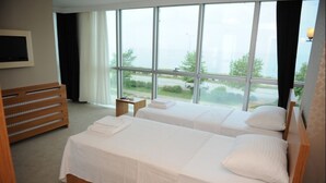 A hotel room with two beds, a wooden dresser, a small table, and large windows offering a view of the outside. - Basoglu Bulancak Otel (Bulancak)