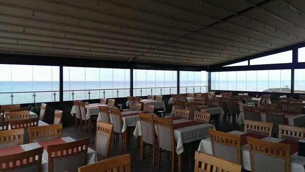 A dining area with tables and chairs, large windows with a view of the sea, and a high ceiling. - Basoglu Bulancak Otel (Bulancak)