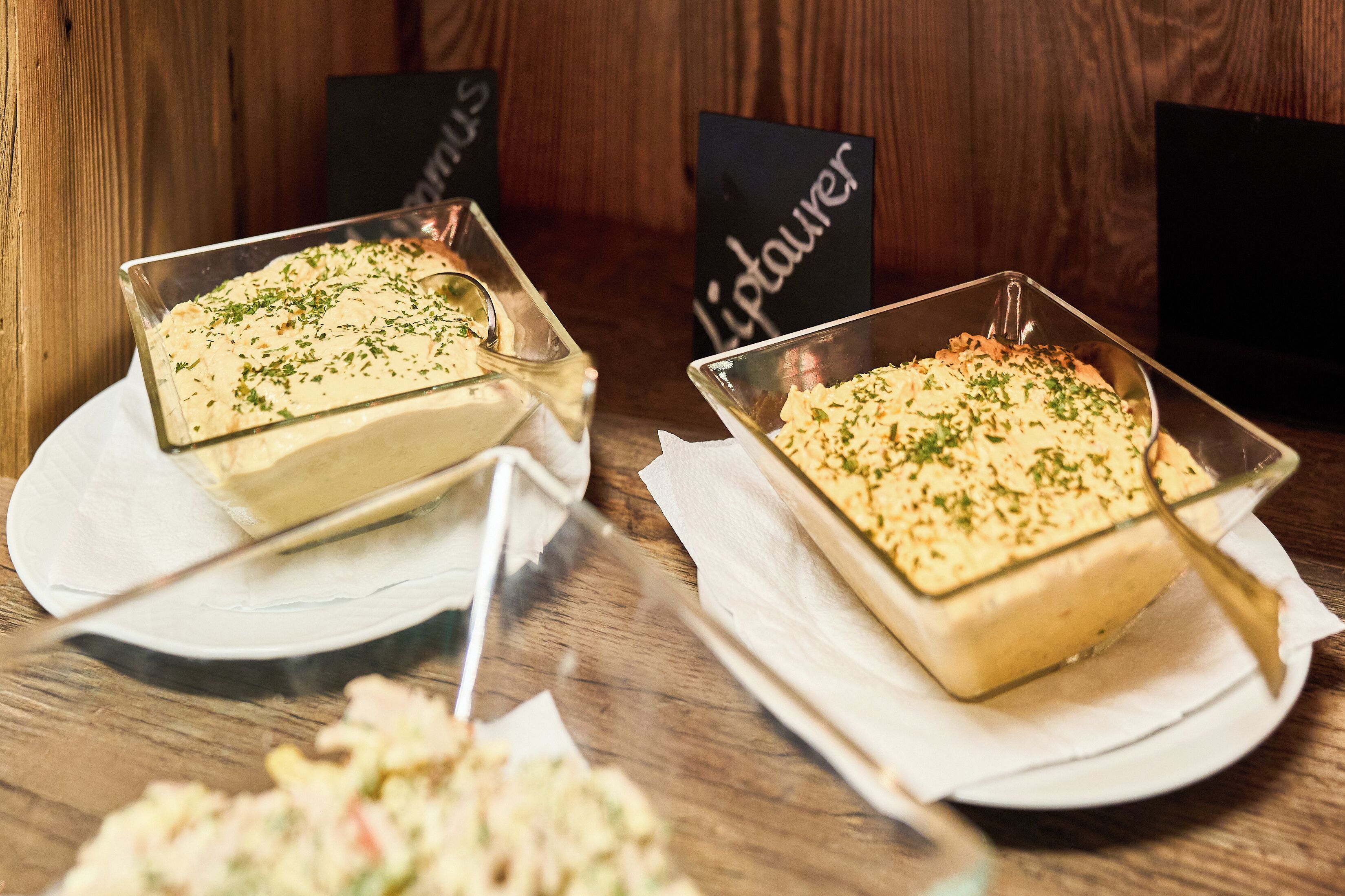 Two square glass dishes of food on white plates with napkins, set on a wooden table.