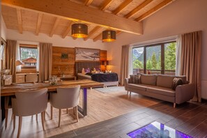 A spacious living area with a wooden ceiling, a dining table with chairs, a sofa, and a large window offering a mountain view. - Hotel Alpennest (Leutasch)
