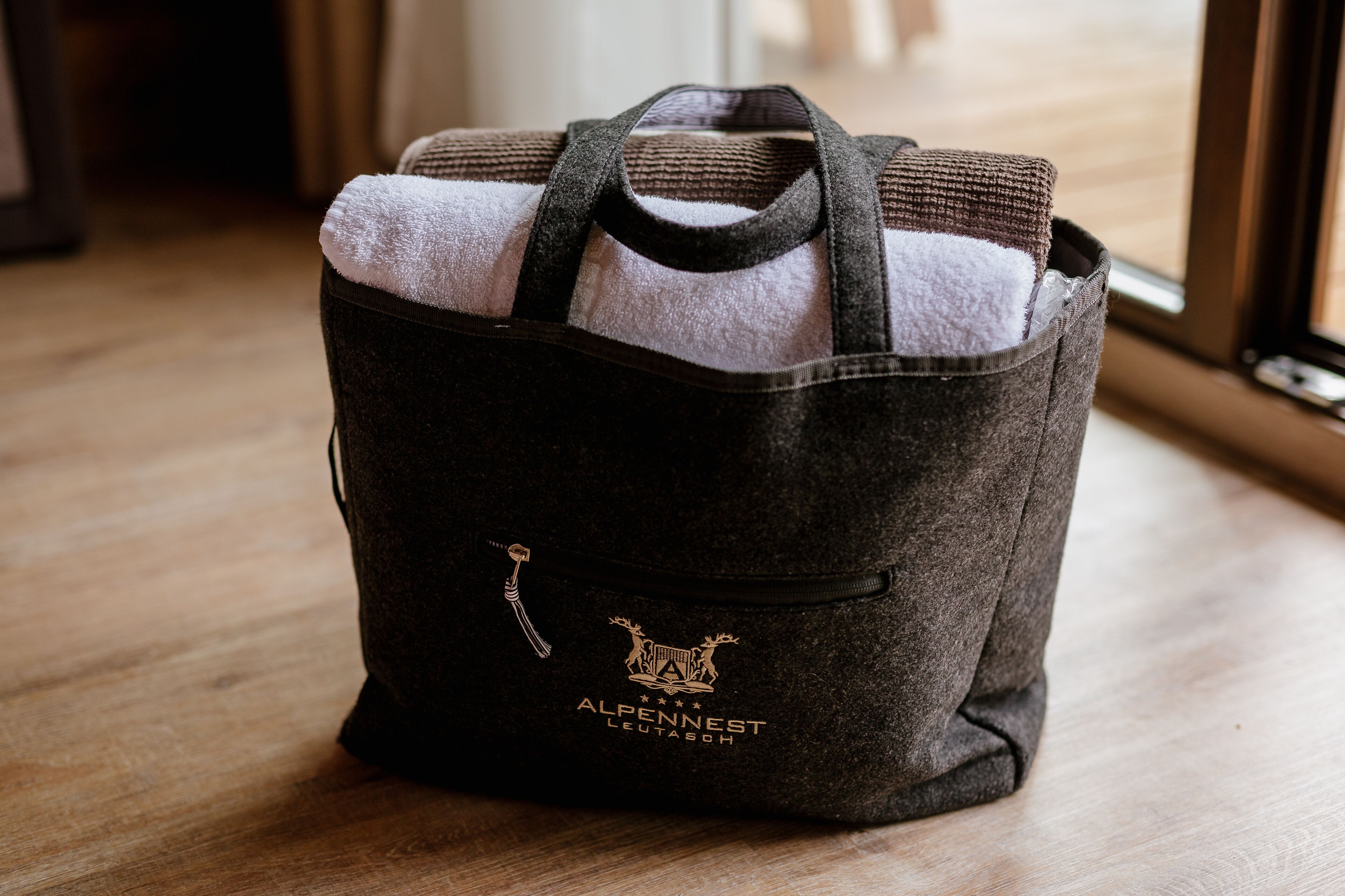 A dark brown tote bag with a logo and text on the front, placed on a wooden floor.