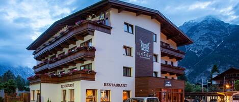 A multi-story building with a restaurant on the ground floor, balconies with flower boxes, and a mountain backdrop.