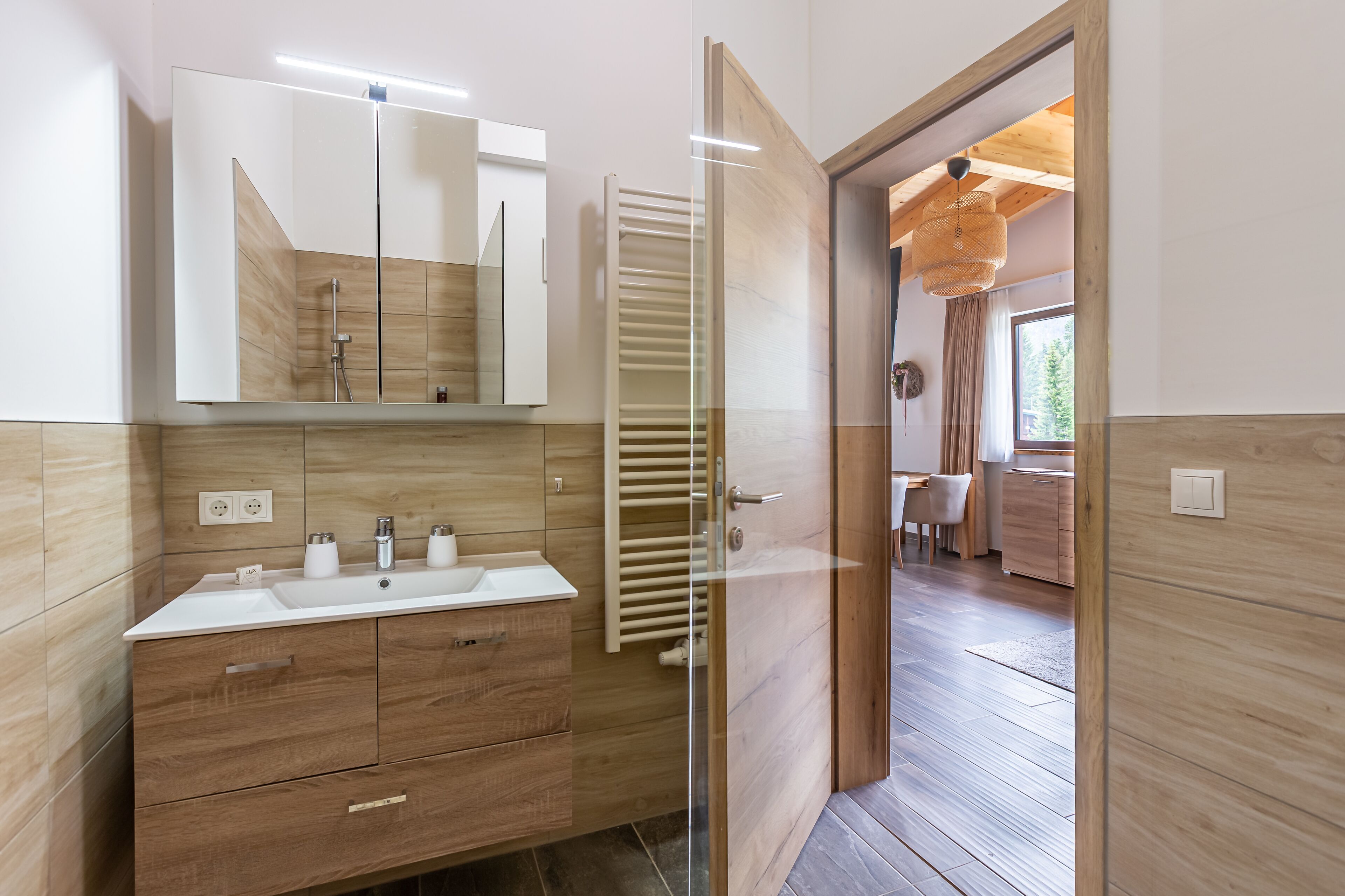 Une salle de bain moderne avec un meuble-lavabo en bois, une cabine de douche en verre et une vue sur l’espace repas.