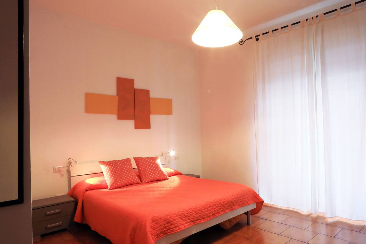 Un dormitorio con una cama, una mesita de noche, una lámpara de pared y una ventana con cortinas.