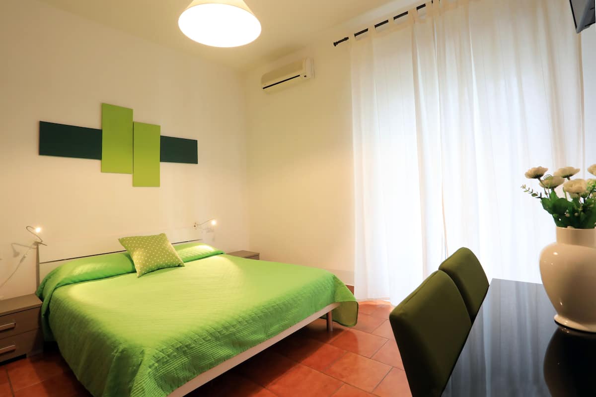 Una habitación con cama, escritorio, silla, aire acondicionado en la pared y ventana con cortinas.
