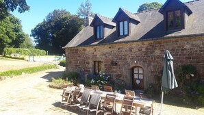 A stone building with a sloped roof, a wooden table with chairs, and a garden area with flowers and plants. - ---------Chestnut - La Châtaigne----------- (Langoëlan)