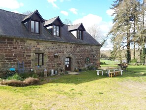 A stone building with a sloped roof, a wooden door, and a small porch area with a bench.