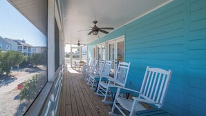 A wooden deck with white rocking chairs and a ceiling fan, overlooking a beach and other buildings.
