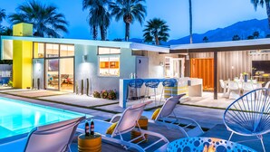 Un'area piscina moderna all'aperto con lettini, un braciere e una cucina visibile attraverso le porte di vetro.