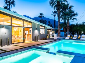 Ett modernt hus med en swimmingpool, glasdörrar som leder till en matplats och palmer i bakgrunden.
