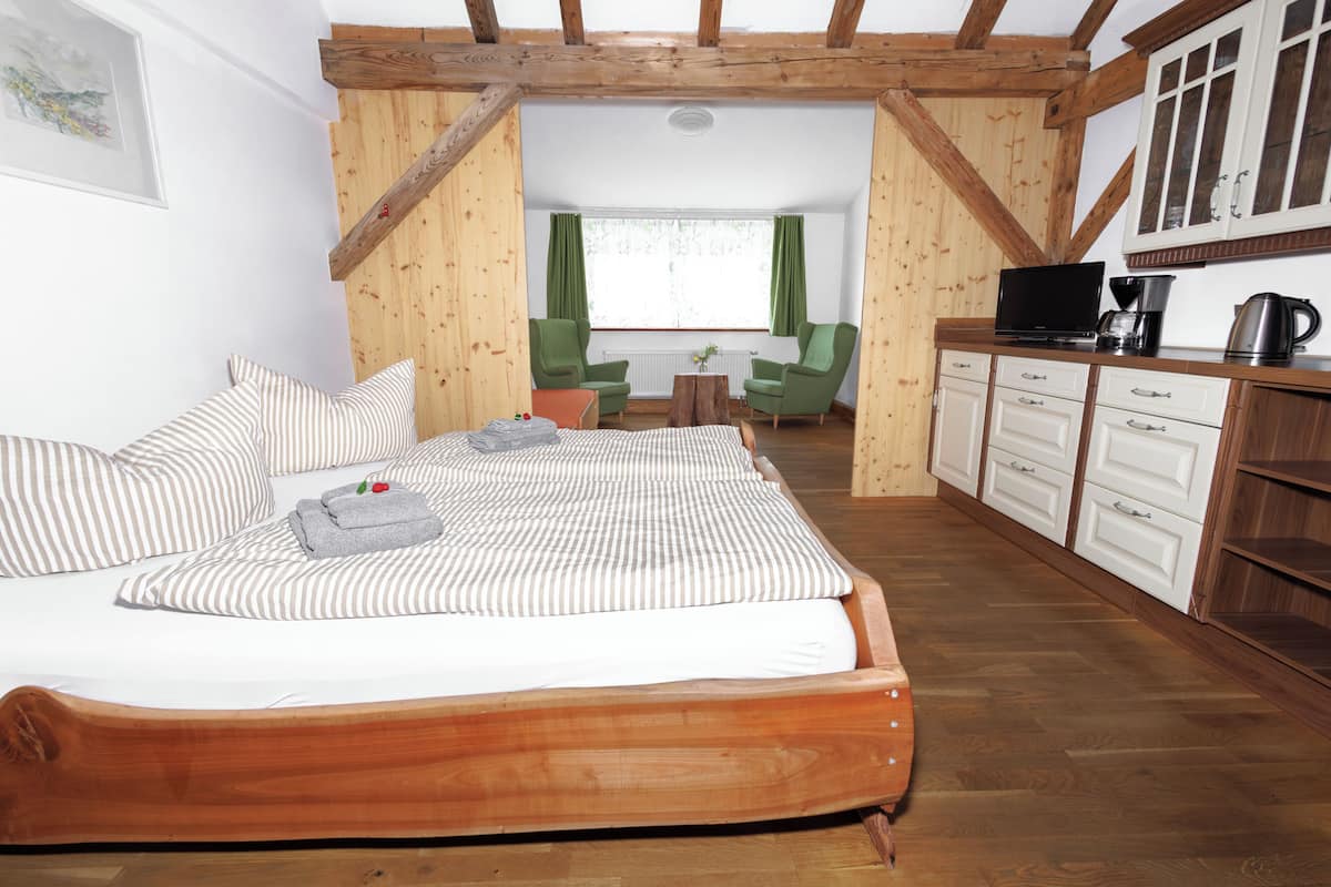 Een slaapkamer met een houten bed, groene stoelen, een kleine tafel en een kitchenette met kastjes en een koffiezetapparaat.