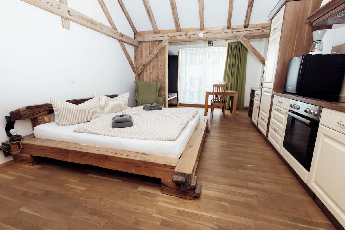 Een slaapkamer met een houten bed, een groene stoel, een klein tafeltje en een kitchenette met een fornuis en koelkast.