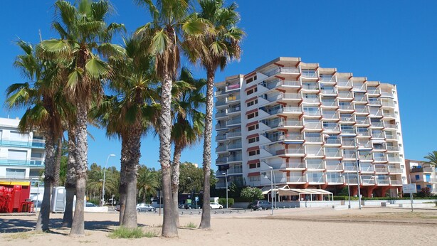 A multi-story hotel with balconies, located near a sandy beach with palm trees.