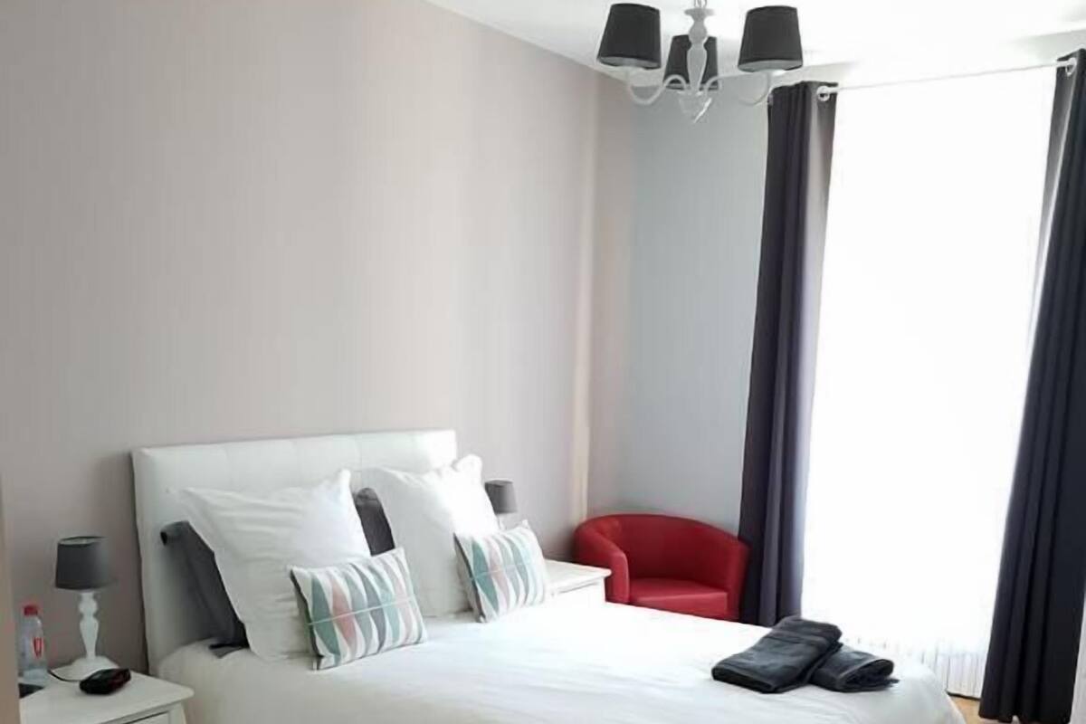 Un dormitorio con una cama grande, una mesita de noche con lámpara, una silla roja y una ventana con cortinas.