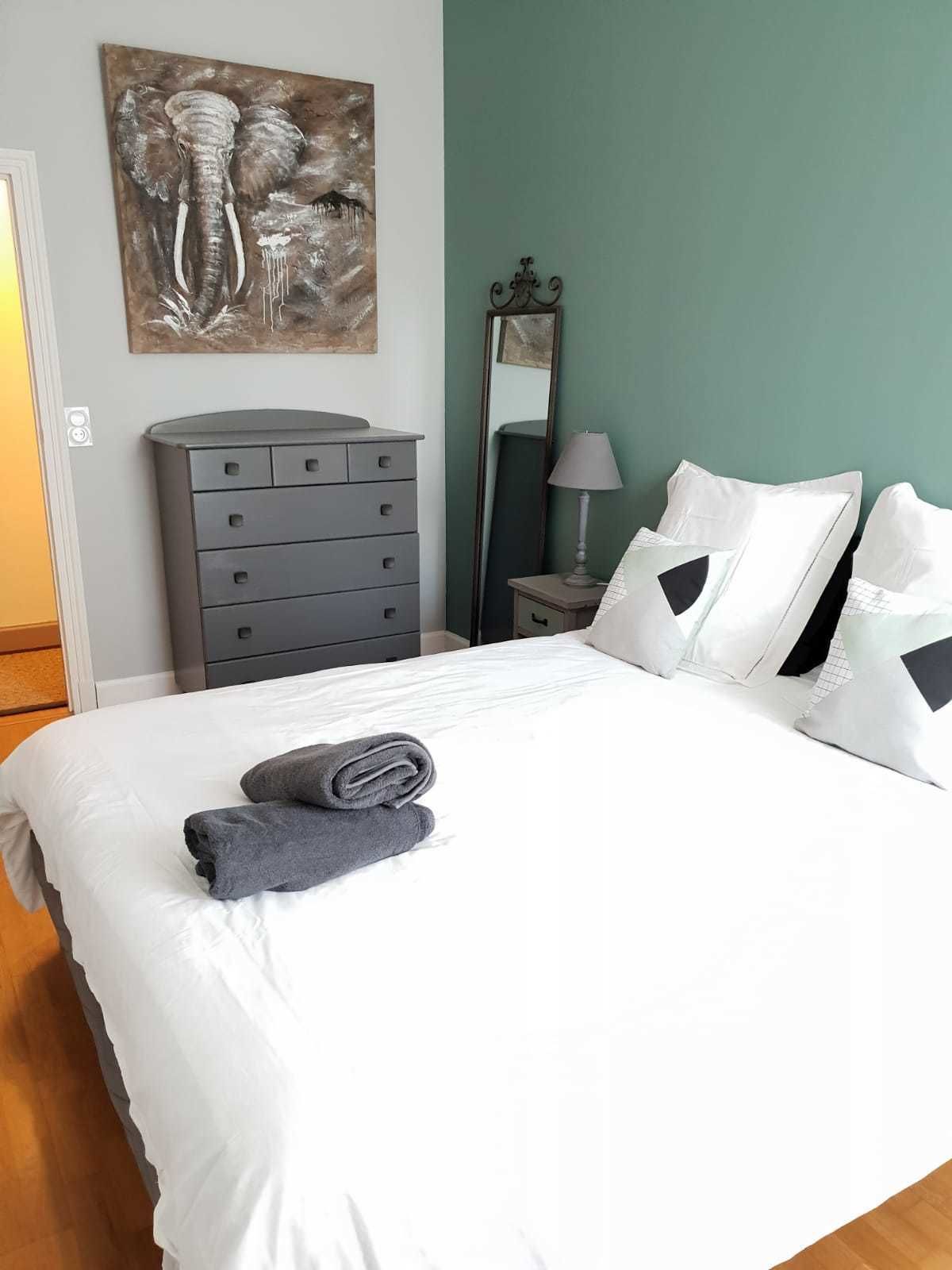 Una cama bien hecha con sábanas blancas y dos almohadas, un aparador gris, un cuadro de elefantes y un espejo en la pared.
