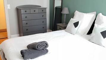 Una cama bien hecha con sábanas blancas y dos almohadas, un aparador gris, un cuadro de elefantes y un espejo en la pared.