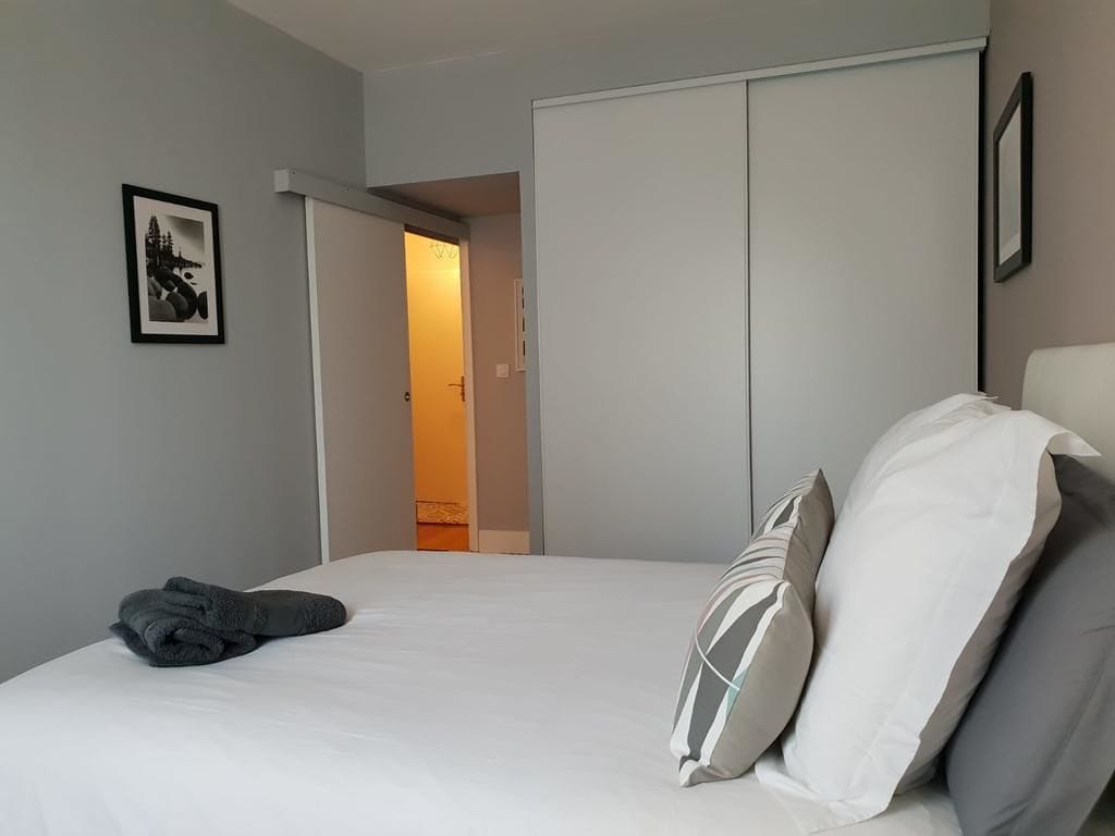 A bedroom with a bed, pillows, a folded towel, a wardrobe, and a door leading to another room.