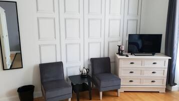 A room with two grey armchairs, a small black table with a teapot, a white dresser with drawers, and a flat-screen TV on top.
