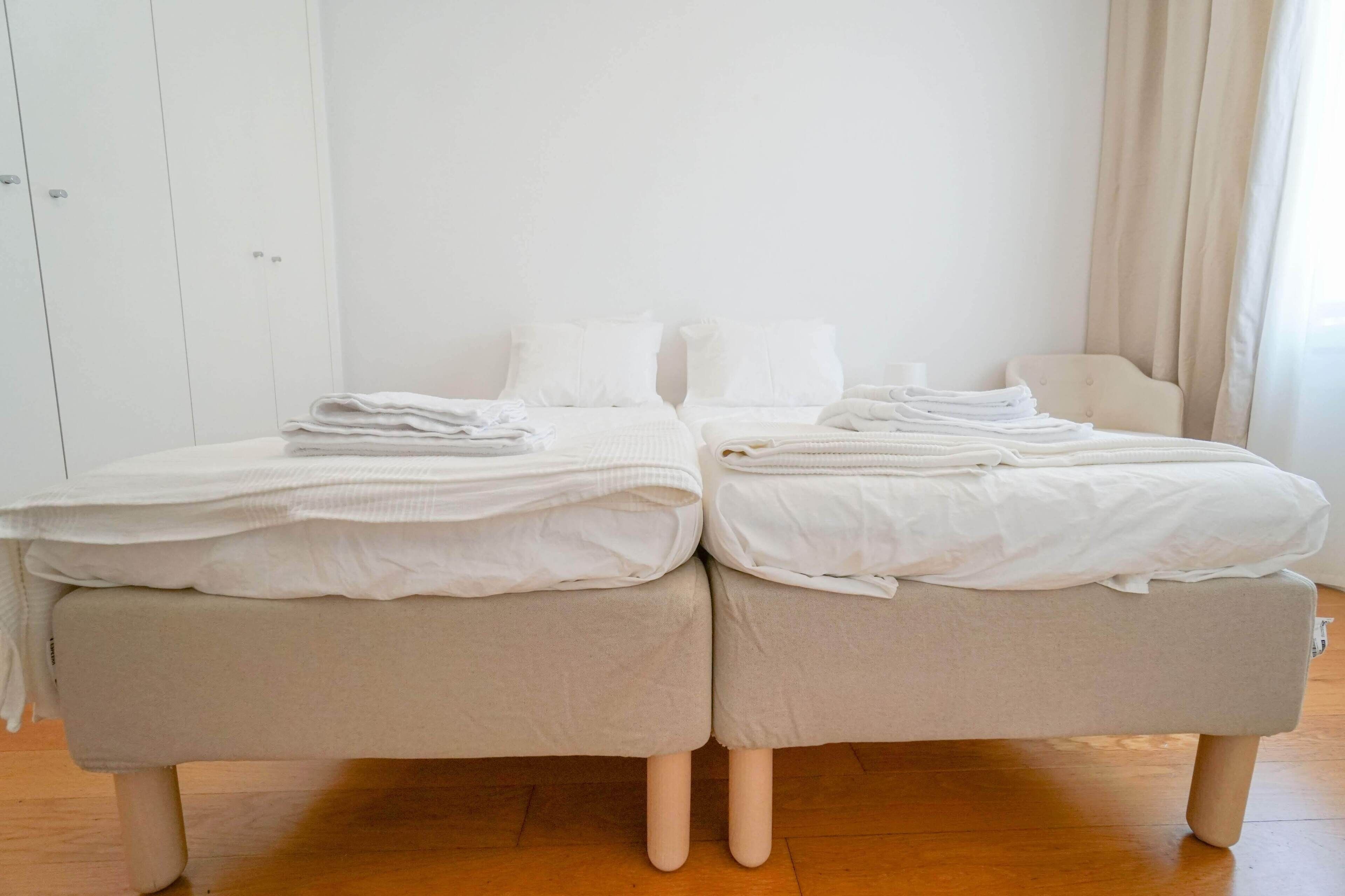 Una cama doble con ropa blanca y un cabecero tapizado en beige, ubicada en una habitación con suelo de madera y paredes blancas.