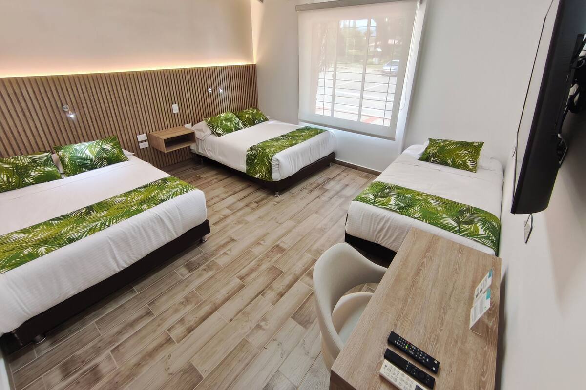 A hotel room with three beds, a wooden floor, a TV, and a window with a view of the outside.