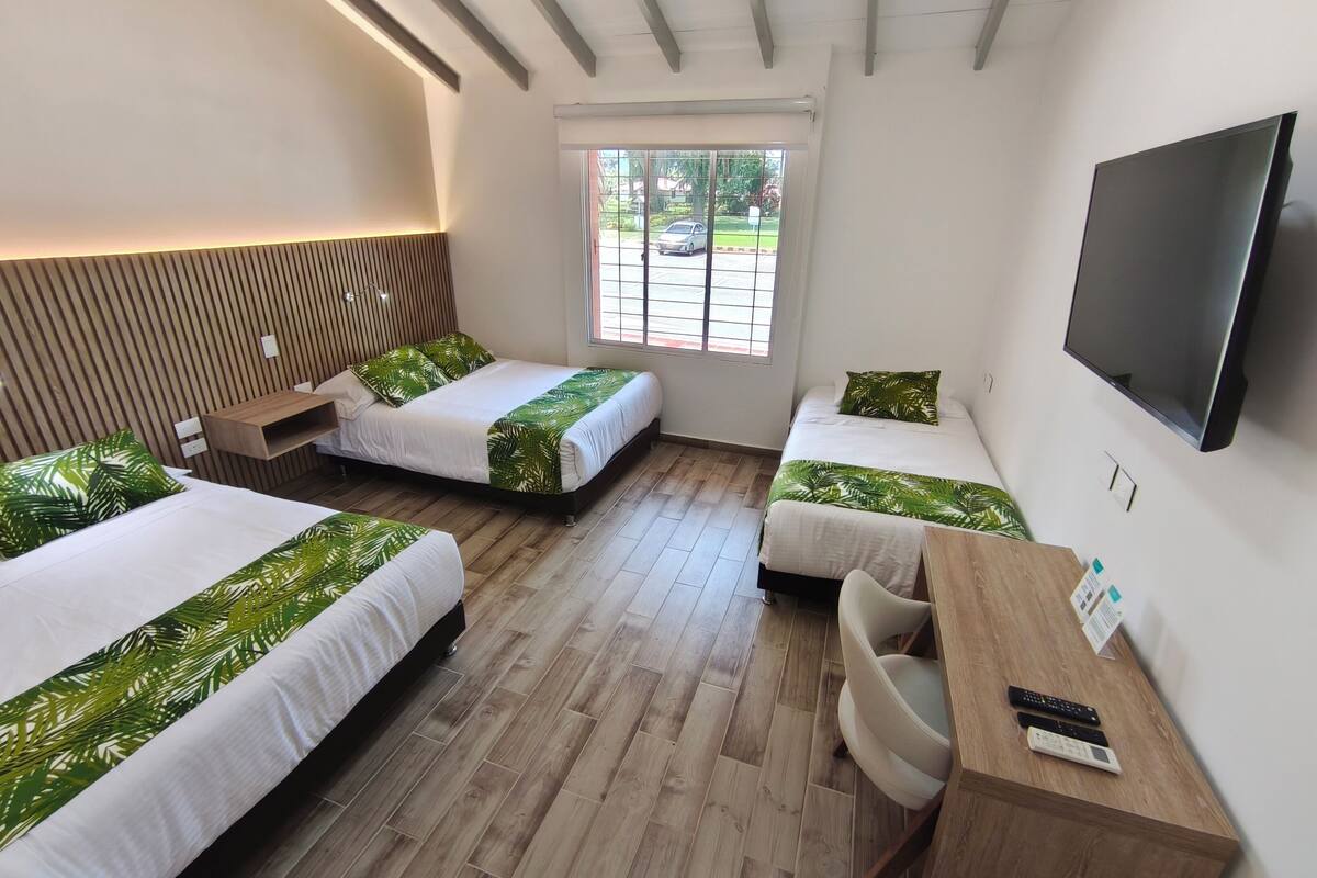 A hotel room with three single beds, a wooden desk, a chair, a television, and a window with a view of the street.
