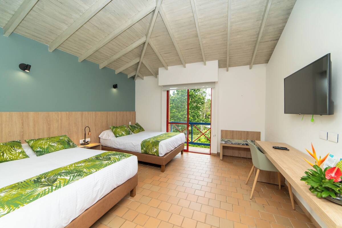 A hotel room with two beds, a desk, a TV, and a balcony with a view of greenery.